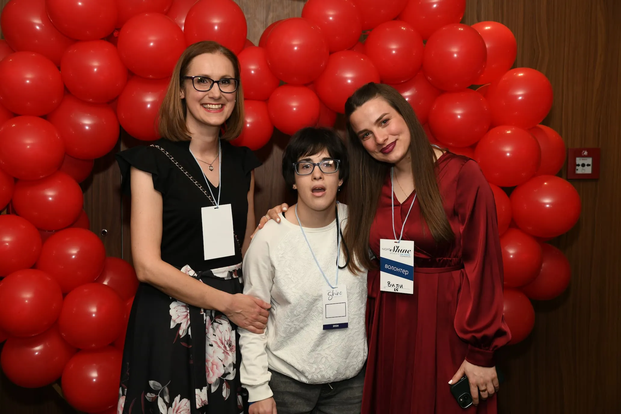 Three friends smiling together in front of a red balloon arch at Night to Shine